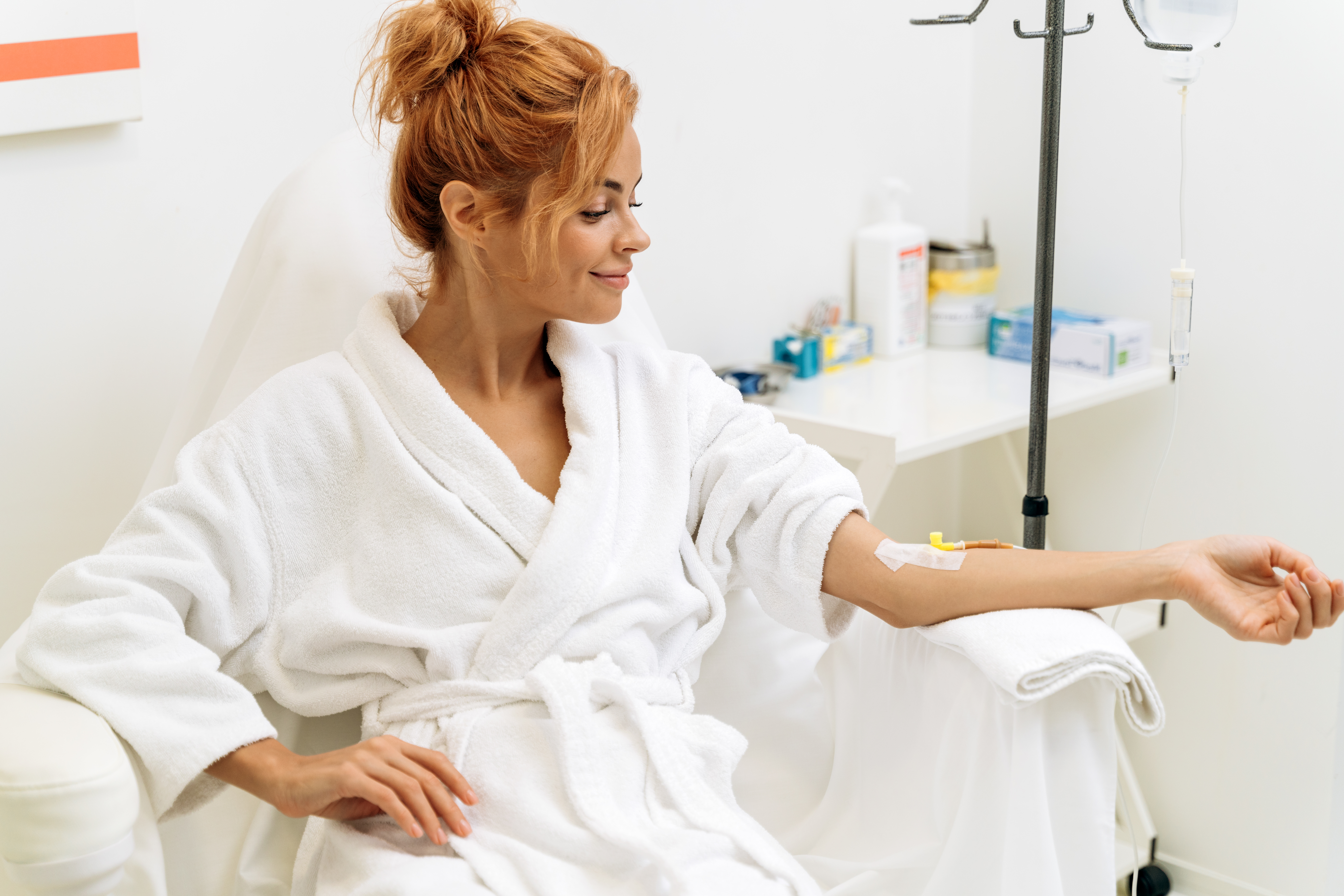 A woman with her hair up in a bun, wearing a white bathrobe, is seated comfortably and smiling while receiving an IV treatment. The IV stand and drip are to her right, and she appears relaxed and content in a clean, well-lit clinical setting.